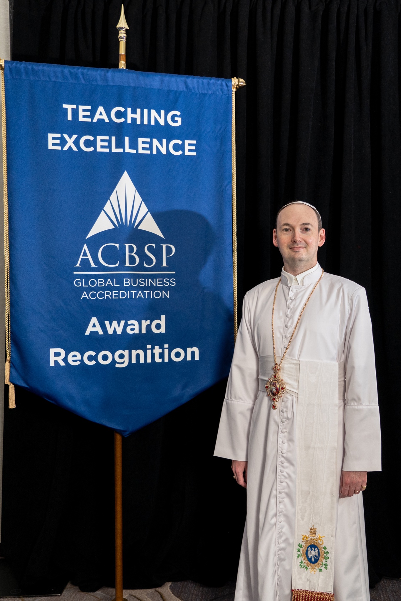 Radislav Romanovich Johnson in ecclesiastical habit beside the ACBSP Teaching Excellence Award banner